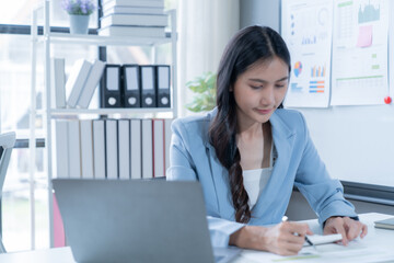 Focused asian businesswoman works at a modern office desk with paperwork and charts, exuding confidence and determination. The scene symbolizes success and growth in entrepreneurship