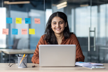 Young woman with laptop in office setting, smiling confidently, showcasing productive and positive workspace. Surrounded by colorful sticky notes representing ideas and planning.