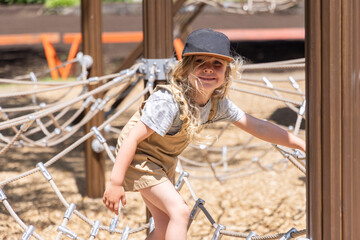 Smiling little boy with long blond hair wearing a cap is playing on a rope bridge at a playground. Selective focus