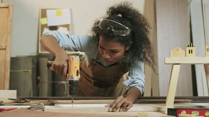 Professional African American female carpenter worker wearing apron standing using electric power drill and screwdriver to assemble by screws to fix into the wooden at workplace in furniture factory. 