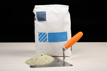 Bag of cement and putty knife on light wooden table against black background. Building material