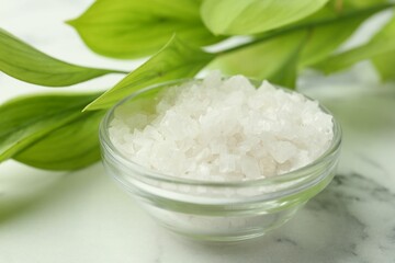 Sea salt in glass bowl and green leaves on white marble table, closeup
