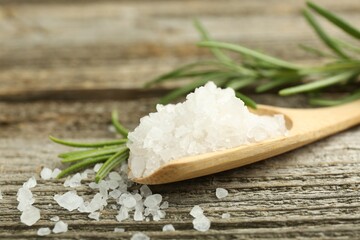 Sea salt in spoon and rosemary on wooden table, closeup