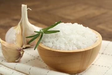 Sea salt in bowl, rosemary and garlic on brown table, closeup