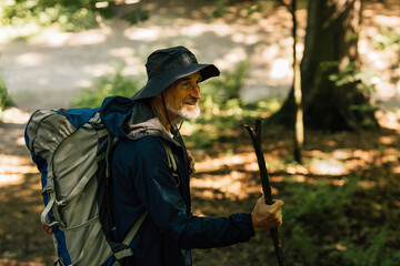Naklejka premium Cheerful senior male with a wooden stick and tourist backpack walking in a forest