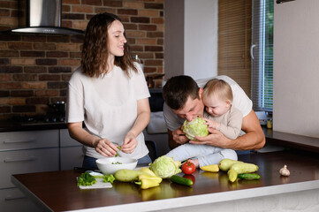 Family in kitchen cooking healthy food, having fun. Child sitting on countertop holding cabbage head, mom, dad, baby taking turns eating it. Healthy eating, organic vegetables, plant-based diet.