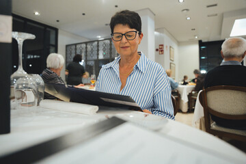 Elegant senior woman is sitting at a restaurant table, carefully reading the menu and deciding what to order