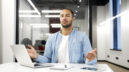 African american businessman meditating with closed eyes during break while sitting at workplace in business office. Calm worker practices yoga and feels peace of mind. Male relaxes to relieve stress
