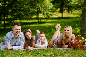 Happy parents with son and daughter lying on stomach during picnic in park, talking to each other, dogs playing nearby. Family weekend concept. Family day