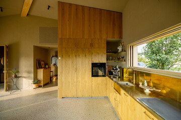 A modern kitchen with golden accents and wooden cabinets, bathed in natural light from the open window. The minimalist design highlights simplicity, while the greenery outside enhances the space