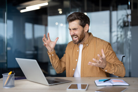 Stressed businessman at modern workspace reacting with frustration. Man sitting at desk with open laptop, notebook, phone, expressing anger or dissatisfaction in office setting.