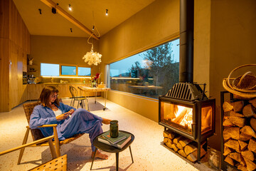 Woman in a blue lounge set sitting by the fireplace, relaxing in a cozy living room with a view of...
