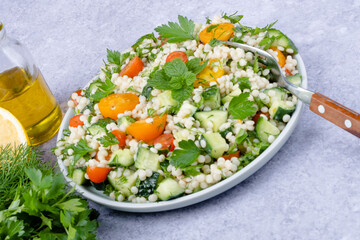 Close up fresh couscous salad with cucumbers, tomatoes, herbs, lemon with a fork on a gray background. Tabbouleh salad. Healthy vegetarian food.