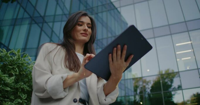 Charming Caucasian woman sitting on bench in small green area next to glass office center. Using tablet device. Using one finger for swiping on touch screen. Selecting items in online store.