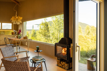 Open kitchen and living area with a fireplace, rustic decor, and a large window overlooking lush greenery. Warm natural light fills the space, creating a peaceful and cozy atmosphere
