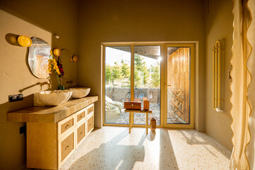 A warm, sunlit bathroom featuring a double vanity with sculpted stone sinks, rattan cabinet details, and a glass door leading to an outdoor area with wooden accents