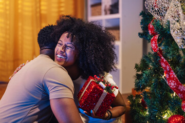 Diverse couple exchanging gifts joyfully near a decorated Christmas tree.