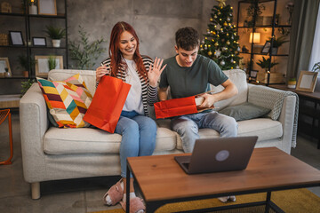 Young cheerful couple open gift from shopping bags during video call