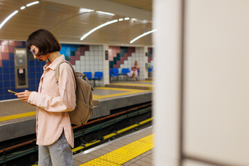 young woman stands at a metro station and looks at her mobile phone. a woman uses a mobile phone at a metro station.