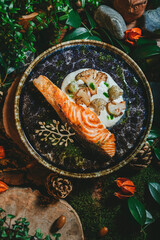 A beautifully arranged plate features a roasted salmon fillet alongside a smooth creamy sauce and thinly sliced vegetables, set in a lush, green backdrop of foliage and decorative elements.