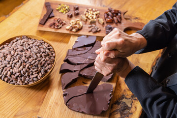 Chocolatier breaking a chocolate bar with a knife, surrounded by ingredients like cocoa beans and nuts. Selective focus