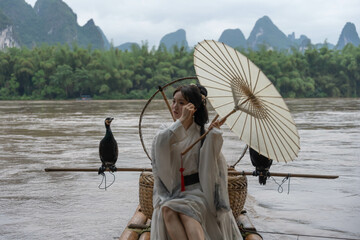 Hanfu girl fixing her hair and holding parasol while standing on bamboo raft by Li river