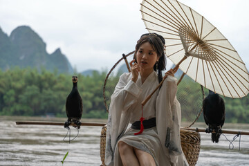 Hanfu girl seated on a bamboo raft holds an umbrella while fixing her hair