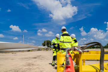 Engineers in high visibility gear assess a large wind turbine blade at a construction site under a bright blue sky, representing the implementation of renewable energy technology for sustainable.