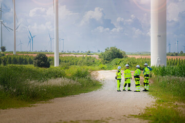 A team of engineers in high-visibility safety gear stand together at a wind farm, conducting...