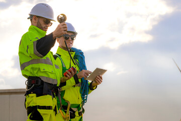Two engineers wearing high visibility safety jackets and helmets measure wind speed with an...