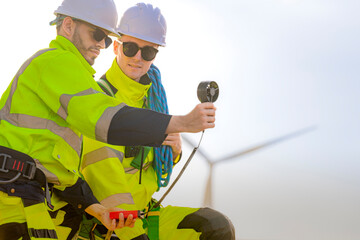 Two engineers wearing high visibility safety jackets and helmets measure wind speed with an...