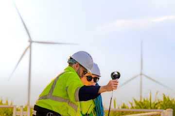 Two engineers wearing high visibility safety jackets and helmets measure wind speed with an...