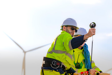 Two engineers wearing high visibility safety jackets and helmets measure wind speed with an...