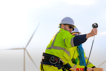 Two engineers wearing high visibility safety jackets and helmets measure wind speed with an...