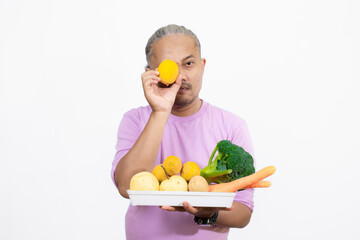 Obraz premium curly-haired Asian adult male carrying a tray of fresh vegetables while pointing at it, healthy and nutritious fresh vegetables eating campaign, healthy lifestyle