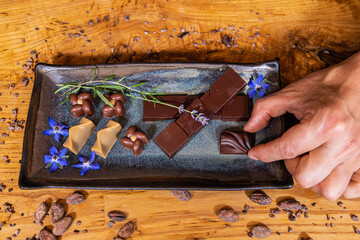 Chocolatier is placing a piece of fine chocolate on a plate, surrounded by other chocolates, flowers, and cocoa beans. Selective focus