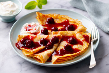Crepes, thin pancakes with cherry, berry sauce and whipped cream. Marble table background. Close up.