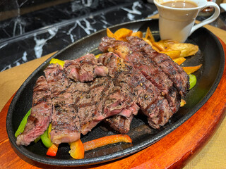 Cast iron plate with sliced grilled rib-eye cooking a medium-rare steak with fried potato and sauce in saucer over table.