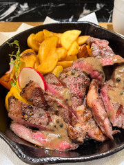 Cast iron plate with sliced grilled rib-eye cooking a medium-rare steak with fried potato and sauce in saucer over table.