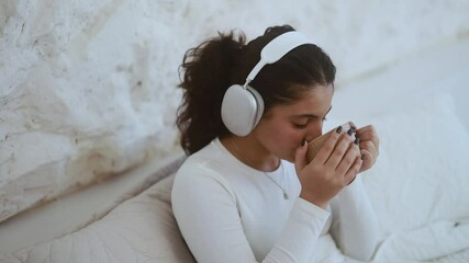 Portrait of charming young indian woman with headphones listening music while laying on bed with cup of hot drink at cozy home bedroom Pretty female enjoying leisure time alone - Powered by Adobe
