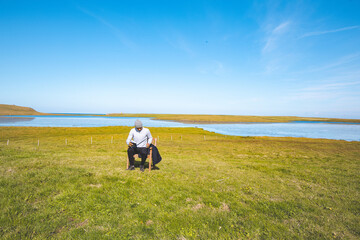 Man sits reading a book in a green field next to a wooden chair, with a serene river and blue sky in the background. The peaceful scene is set in the tranquil landscape of western Iceland