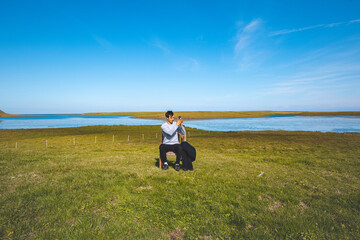 Man sits on a wooden chair in a vast green field, taking a selfie with his phone against the backdrop of a calm river and blue sky in western Iceland. Deep connection with nature