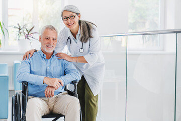Portrait of smiling adult professional therapist embracing shoulders of older disabled happy male...