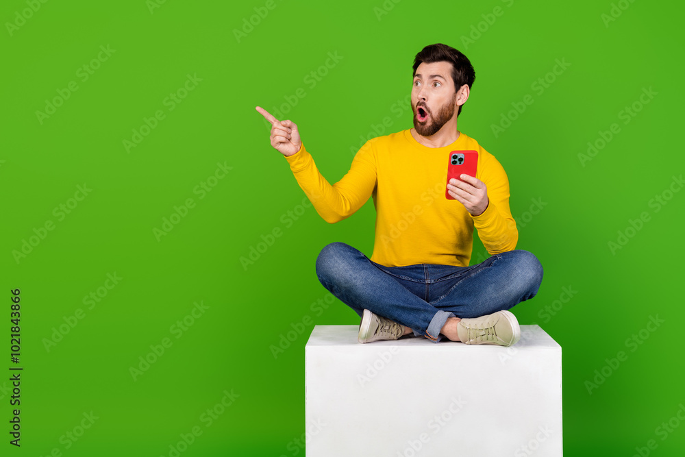 Poster full length photo of handsome guy point look amazed empty space sit cube gadget dressed yellow garme