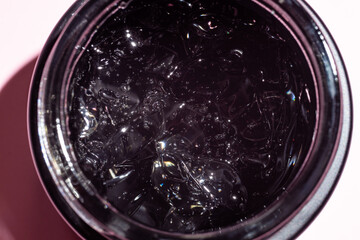Jar with transparent cosmetic product, gel or cream in black glass jar on pink colored background. Unbranded package with of antioxidant cream for self-care. View from above.