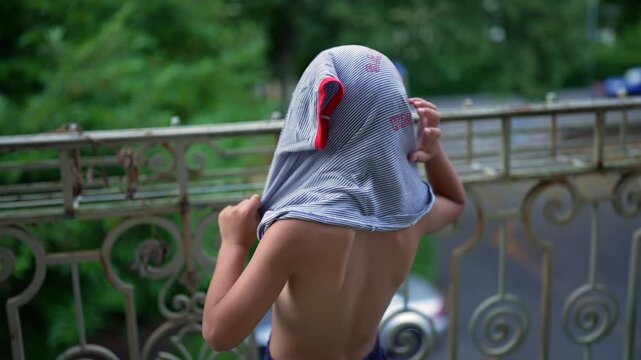 Young boy putting on a shirt while standing on a balcony, looking out into the world, moment of contemplation and readiness, peaceful outdoor scene in a residential area