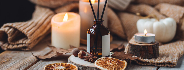 Hello autumn, cozy slow living. Pumpkin, aromatic candle, warm sweaters, dry fall leaves, cinnamon, anise in golden lights with bokeh on white wooden background. Thanksgiving apartment decor banner