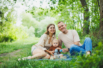Fototapeta premium A family of a mother and father with a child in their arms, sitting in the park in the summer. The concept of a happy family with a small child, care and love. Happy family with a child.