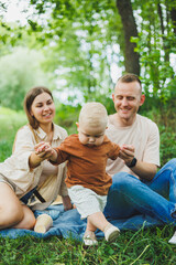Fototapeta premium A family of a mother and father with a child in their arms, sitting in the park in the summer. The concept of a happy family with a small child, care and love. Happy family with a child.