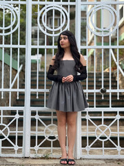 Beautiful brunette girl posing in sexy and fashionable clothes in front of white old-style iron gate. Full length shot outdoors in daylight